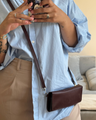 Person wearing a light blue shirt and beige pants, holding a phone with a floral case and carrying markore gul sahar crossbody bag handmade from full-grain leather