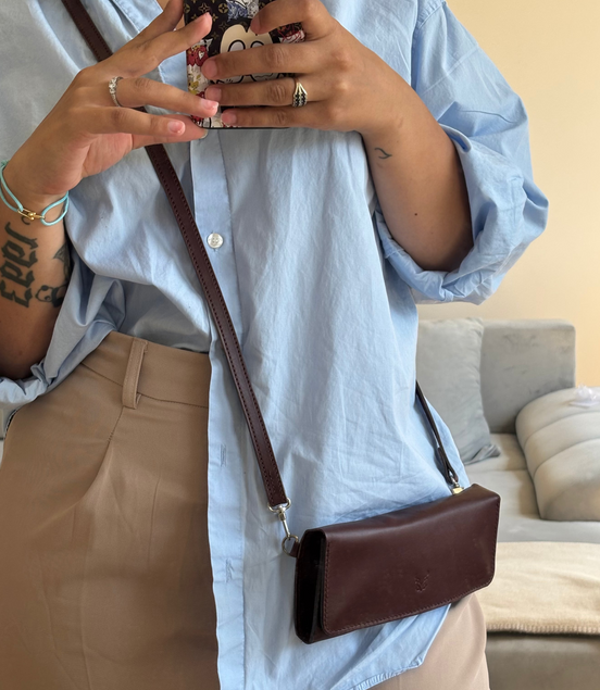 Person wearing a light blue shirt and beige pants, holding a phone with a floral case and carrying markore gul sahar crossbody bag handmade from full-grain leather