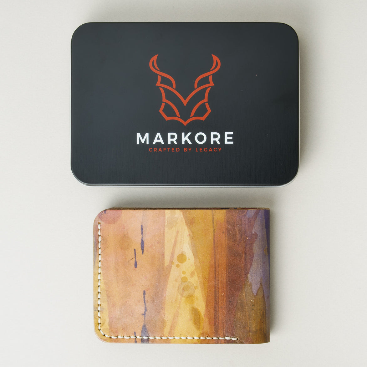 Hand-painted full-grain leather wallet with abstract art in purple, amber, and brown, displayed below a branded packaging tin. Markore Canvas Chaos.