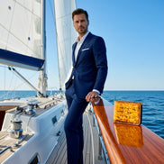 Full-body view of a man in a navy suit standing on the deck of a modern sailing yacht, with Markore Canvas Flame in Solar Flare placed prominently on the polished wooden railing in the foreground. White sails and the open sea form the background, giving a premium maritime lifestyle feel.