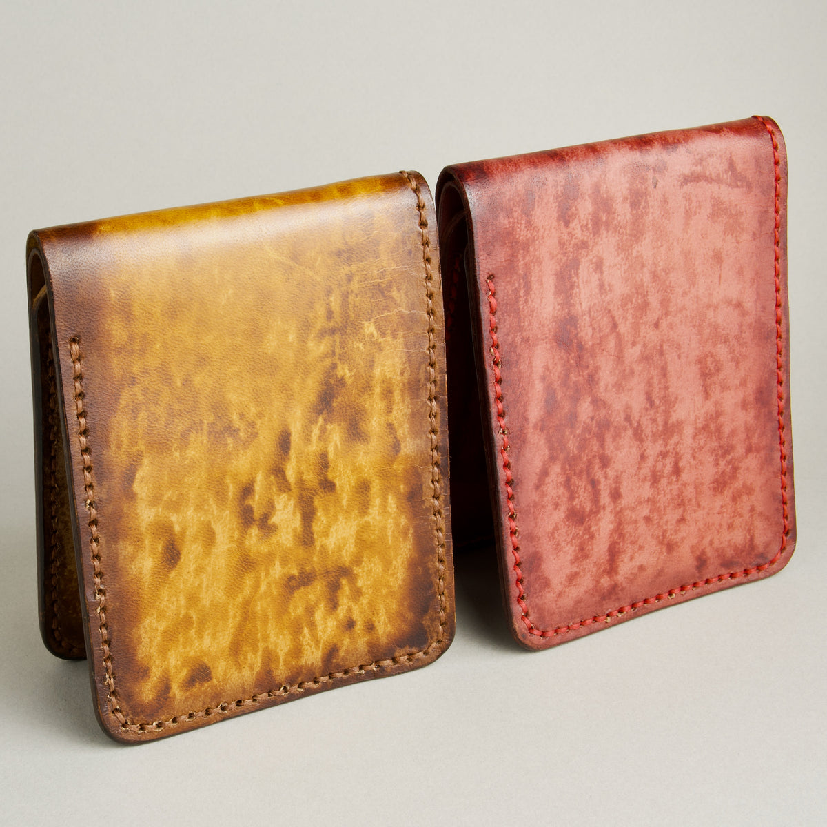 Two of hand-painted full-grain leather bifold wallets. One in amber and brown and one in red, featuring handmade stitching. Markore Canvas Flame.