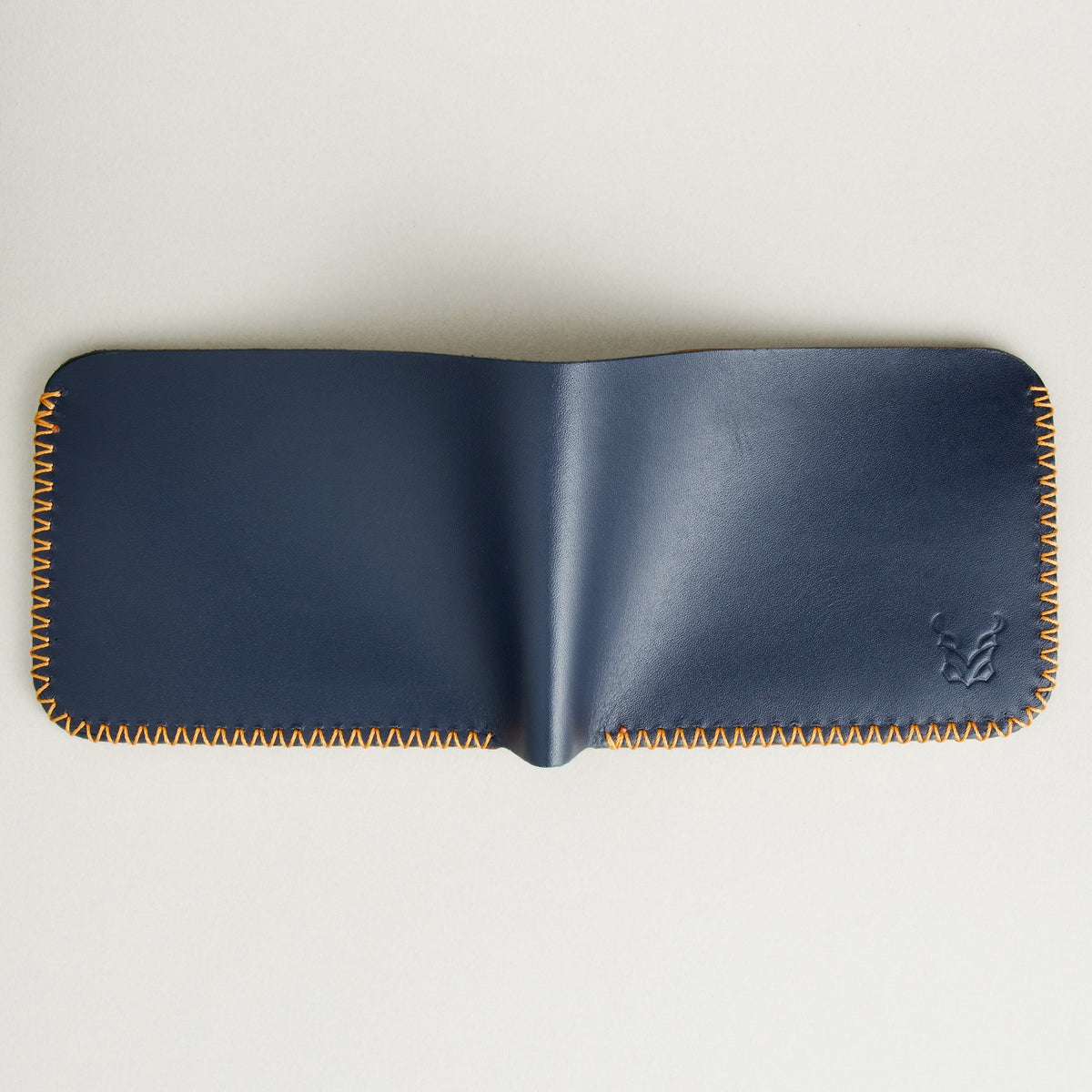 Back view of unfolded dark blue full-grain leather bifold wallet featuring handmade amber cross-stitched detailing on the edges. Markore Hunza Edge.