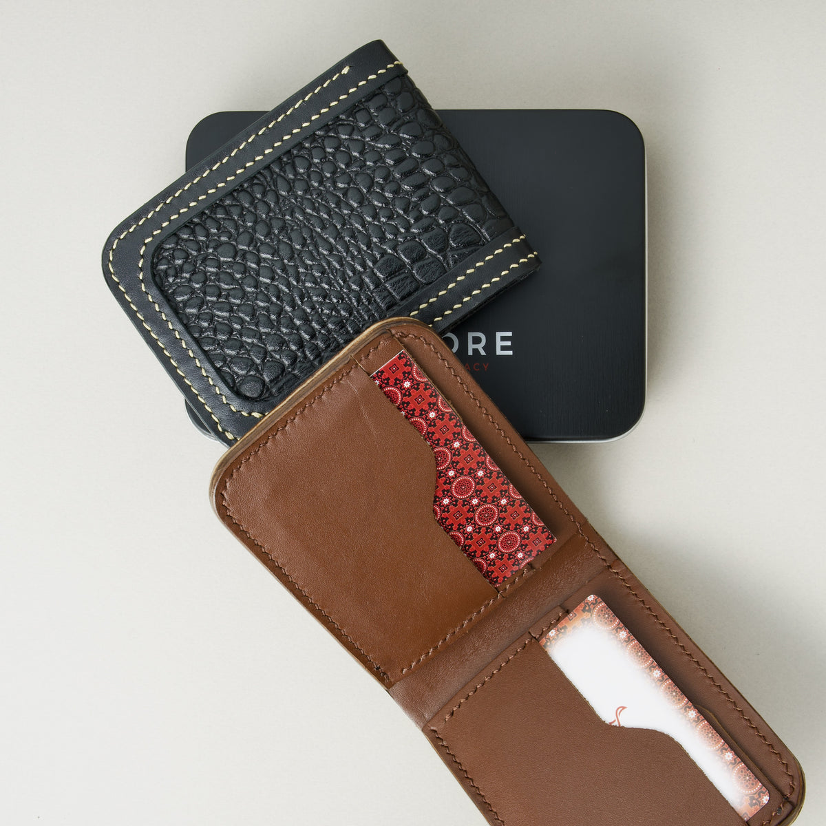Front view of obsidian black croc-embossed full-grain leather bifold wallet and interior view of espresso brown wallet placed on a black Markore branded box.
