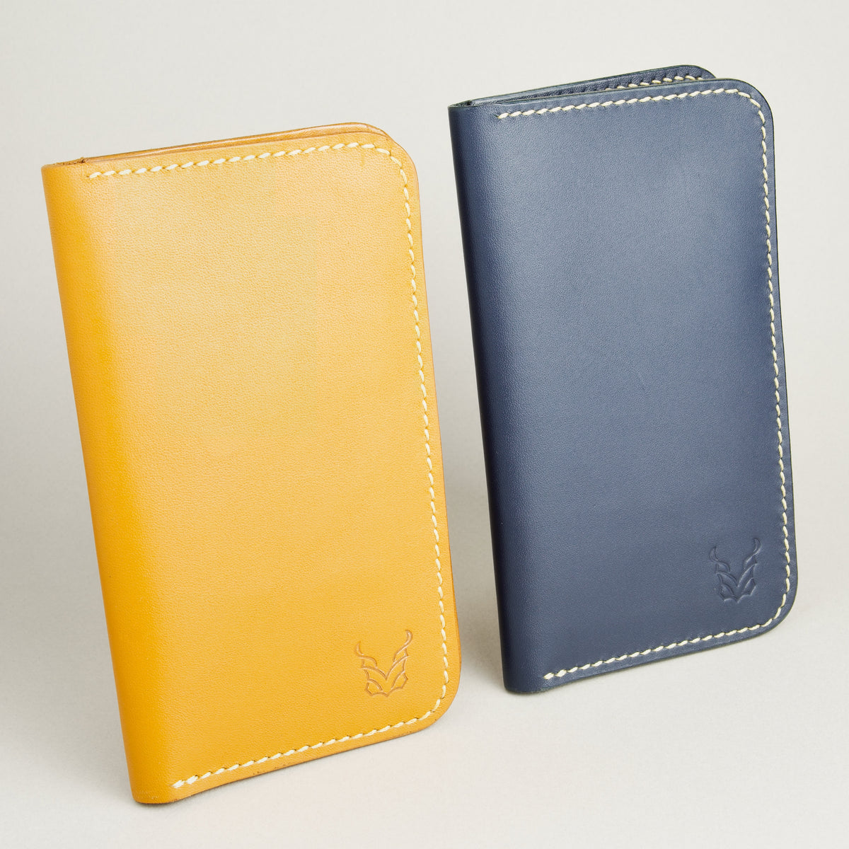 Side-by-side view of two vertical full-grain leather bifold wallets in Amber and Dark Blue, featuring handmade stitching and embossed logo. Markore Karakoram II.