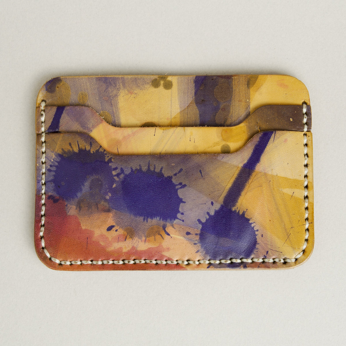 Backside view of hand-painted full-grain leather card sleeve in purple, amber, and brown, featuring handmade stitching. Markore Niva Chaos.