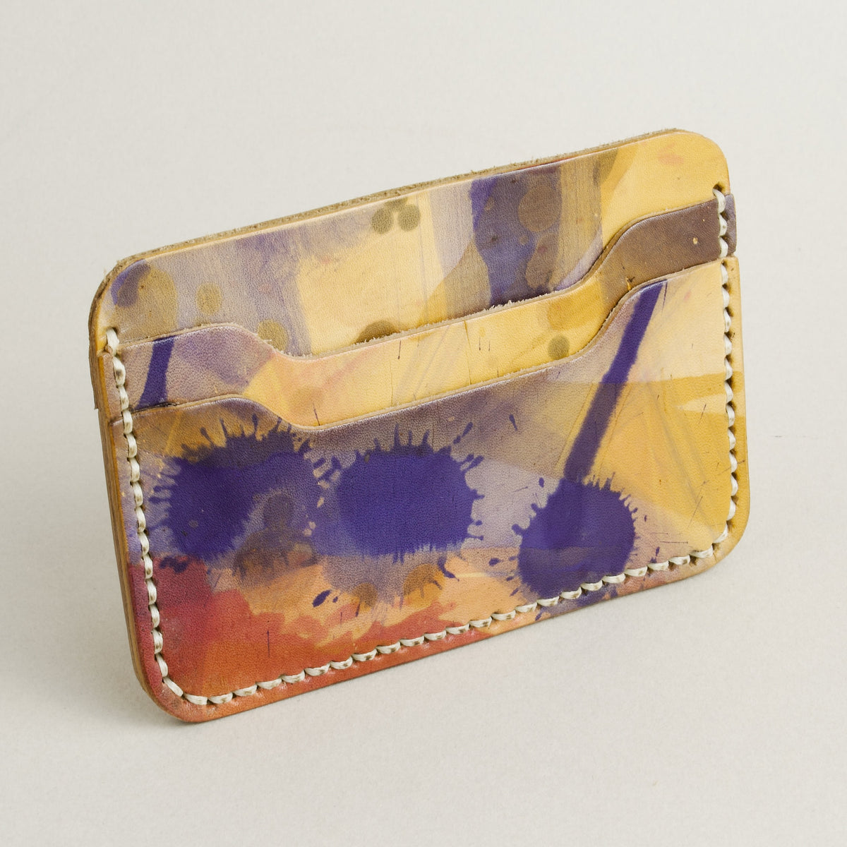 Side view of hand-painted full-grain leather card sleeve in purple, amber, and brown, featuring handmade stitching. Markore Niva Chaos.