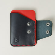 Front view of full grain leather card holder in black and red featuring an open side flap with two stainless steel press buttons, red handmade stitching and embossed logo. Markore Niva Glide.