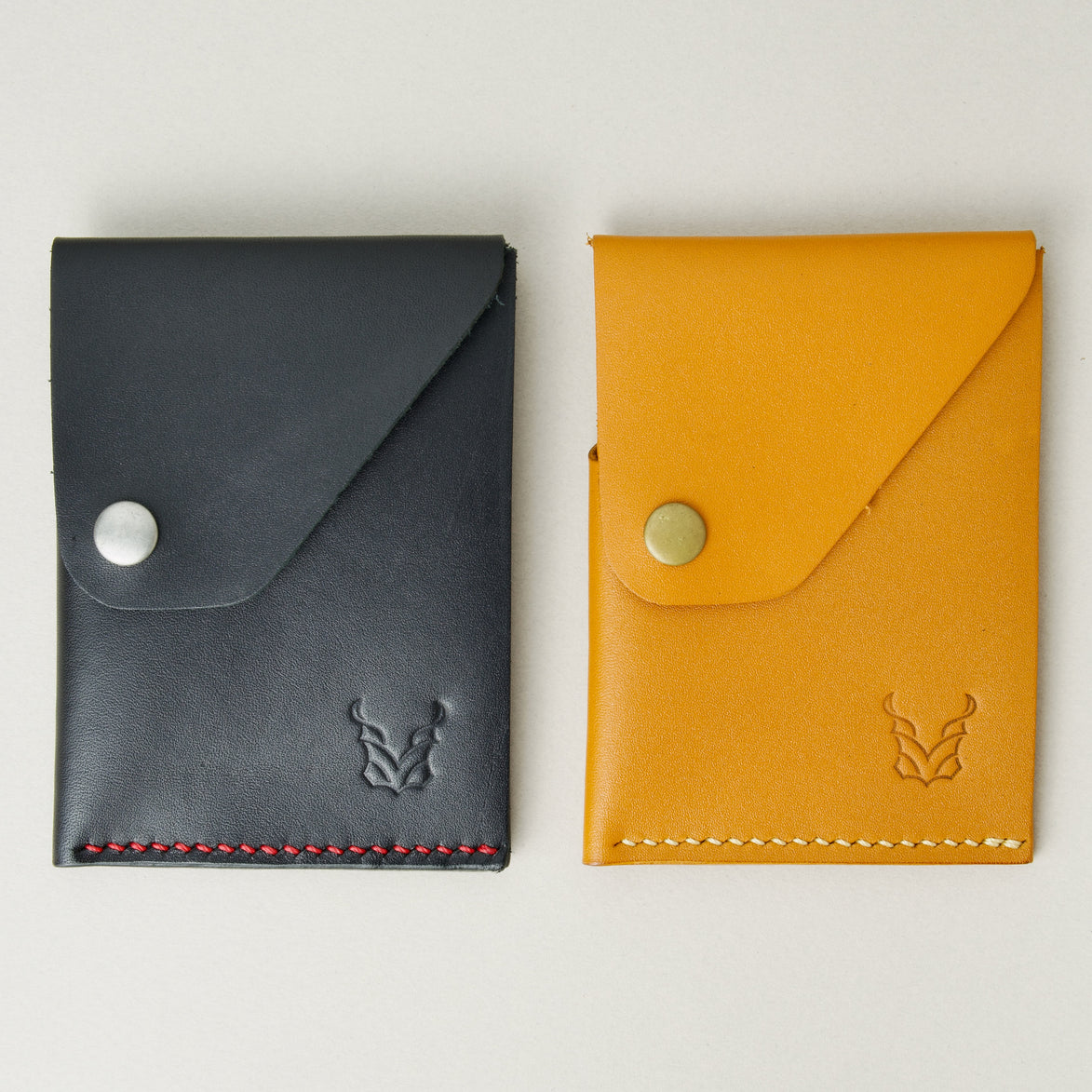Side by side view of two full grain leather wallets in midnight black and amber, featuring handmade stitching, a flap with a press button and embossed logo. Markore Niva Heritage.