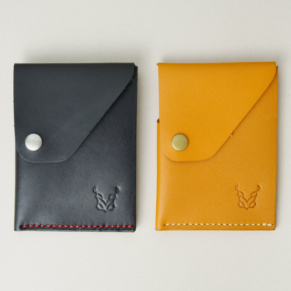 Side by side view of two full grain leather wallets in midnight black and amber, featuring handmade stitching, a flap with a press button and embossed logo. Markore Niva Heritage.
