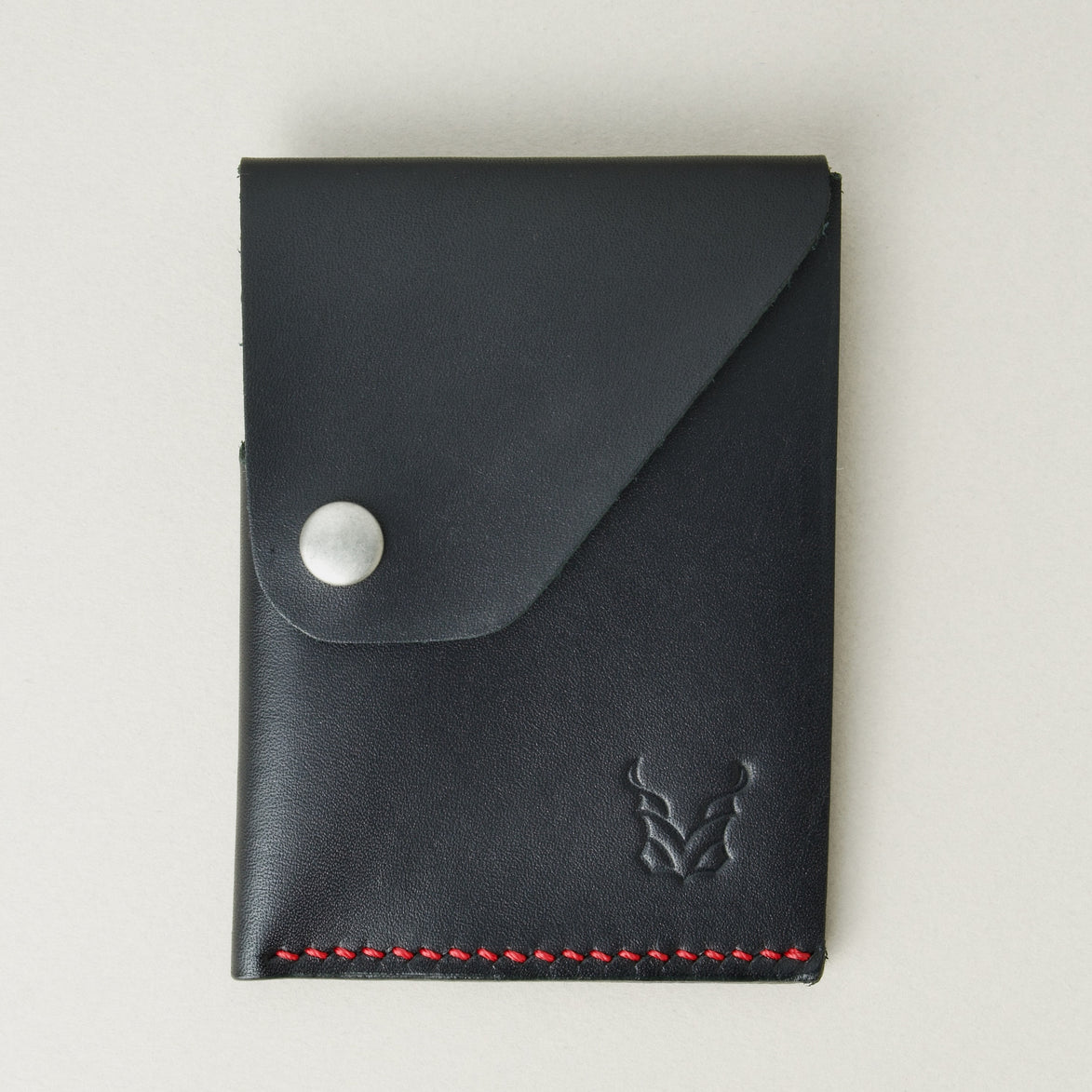 Front view of full grain leather card holder in black with red handmade stitching, featuring a flap with stainless steel press button and embossed logo. Markore Niva Heritage.