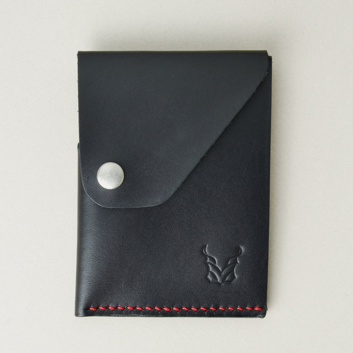 Front view of full grain leather card holder in black with red handmade stitching, featuring a flap with stainless steel press button and embossed logo. Markore Niva Heritage.