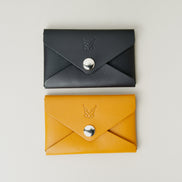Side-by-side view of two full grain envelope style wallets in midnight black and amber with stainless steel press button and embossed logo. Markore Niva Seal