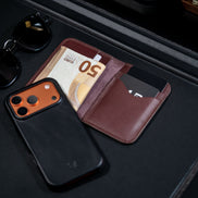 markore ravi slip bifold wallet in espresso brown with cash in it and markore shell frame iphone 17 pro leather case sitting on a black surface.